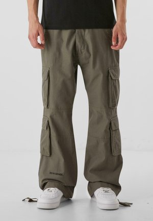 Osoba ubrana w oliwkowe spodnie typu cargo z wieloma kieszeniami i białe trampki, stojąca na jednolitym tle.