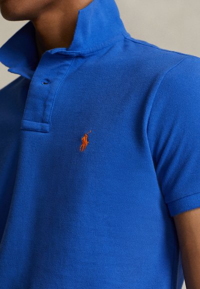 Μπλε πόλο πουκάμισο με υφασμάτινη υφή, διπλωμένο γιακά και πορτοκαλί κεντητό λογότυπο στο αριστερό στήθος. Κοντά μανίκια και εφαρμοστή γραμμή.