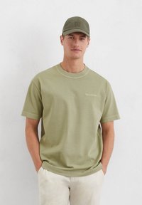 Olive green cotton t-shirt with short sleeves, crew neckline, and subtle logo detail. Paired with a matching cap. Smooth texture.