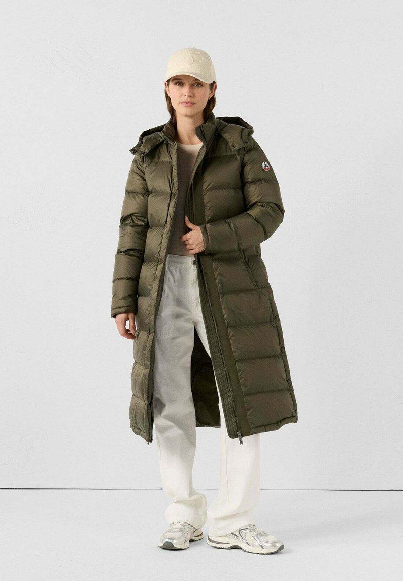 Cappotto imbottito lungo di colore verde oliva con cappuccio, caratterizzato da cuciture verticali e zip, abbinato a pantaloni crema e sneaker.