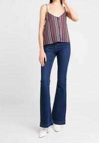 Camisole rayée multicolore avec des fines bretelles et des boutons sur le devant, associée à un jean évasé bleu foncé et des chaussures à bouts pointus de couleur claire.