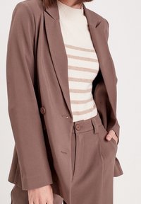 Blazer marron ajusté avec un design croisé, de larges revers et deux boutons. Associé à un haut à rayures crème et un pantalon assorti.