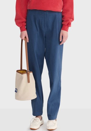 Person wearing blue high-waist pants, red long-sleeve top, white sneakers, holding beige bucket bag with brown strap by their side.