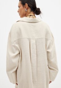 Camicia in lino beige con vestibilità ampia, dotata di colletto, maniche lunghe e dettaglio a piega sulla schiena. Texture liscia con una leggera lucentezza.