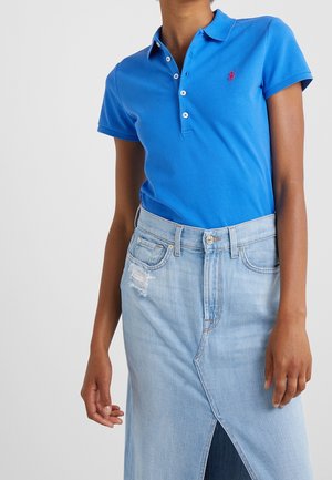 Polo azul de manga corta con logo rojo, metido dentro de una falda vaquera de tiro alto en azul claro, con abertura frontal y parche desgastado.