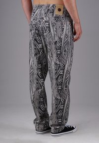 Man wearing black and white patterned loose-fit pants and black sneakers with white soles, standing against a plain gray background.