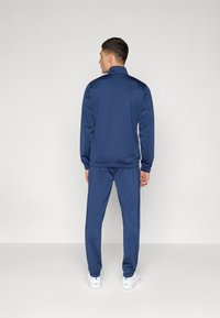 Navy tracksuit made of smooth fabric. Features a zip-up collar, elastic cuffs, and matching pants with a tapered fit. White sneakers complete the look.