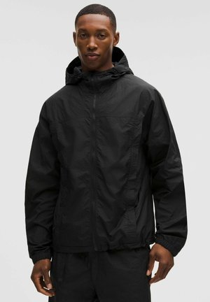 Nuori mies, joka pukeutunut mustaan vetoketjulliseen hupputuulipusakkaan ja siihen sopiviin mustiin housuihin, yksinkertaisen vaalean taustan edessä.