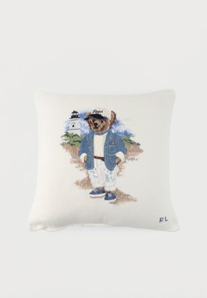 Balta pagalvėlė su išsiuvinėta meška, dėvinčia džinsinę striukę, baltas kelnes, mėlynas batus ir kepurę, stovinti prie švyturio ant smėlio fono.