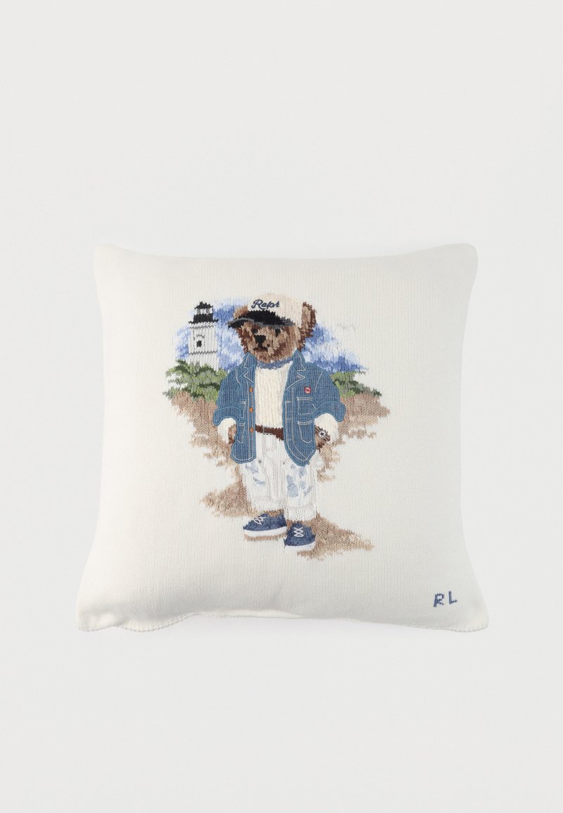 Λευκό μαξιλάρι με κεντημένη αρκούδα που φοράει τζιν μπουφάν, λευκό παντελόνι, μπλε παπούτσια και καπέλο, στέκεται κοντά σε φάρο σε αμμώδες φόντο.