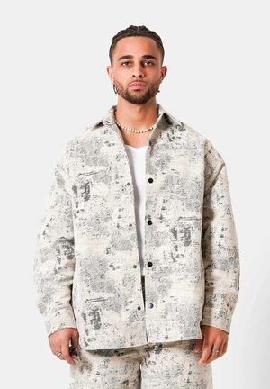 Junger Mann mit geflochtenem Haar trägt eine gemusterte beige Jacke und passende Hose über einem weißen Hemd, steht vor einem schlichten hellen Hintergrund.