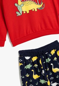 Sudadera roja con un gráfico de dinosaurio, combinada con pantalones cortos marinos que presentan varios patrones de dinosaurios en amarillo, blanco y verde.