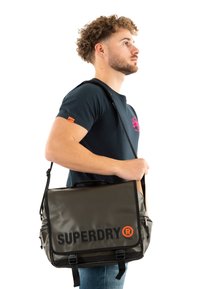 Sacoche grise avec rabat, présentant un grand logo noir "SUPERDRY" et un accent orange. Fabriqué en matériau texturé, il comprend une bandoulière réglable.
