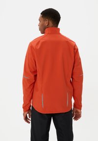 Rote Softshell-Jacke mit hohem Kragen, elastischen Bündchen und reflektierenden Akzenten. Sie verfügt über ein schlankes Design und eine glatte Oberfläche.