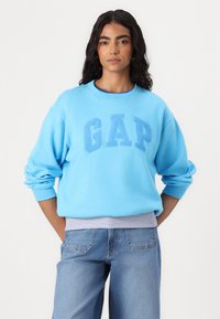 Sudadera azul clara con un logo en relieve de "GAP", cuello redondo y puños acanalados, combinada con jeans azules claros y cinturilla a rayas.