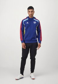 Fato de treino Adidas com um capuz azul com detalhes em vermelho, amarelo e branco, combinado com calças pretas e chuteiras de futebol brancas.