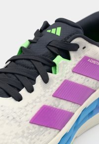 Scarpa sportiva bianca con dettagli viola e blu, caratterizzata da una tomaia testurizzata, lacci neri e un logo verde.