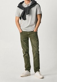 Polo grigio, jeans verde oliva e sneakers bianchi. Un felpa nera è appoggiata sulle spalle. Outfit semplice e casual con colori neutri.