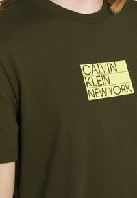 Olivově zelené bavlněné tričko s kulatým výstřihem a krátkým rukávem. Na přední straně je žlutý grafický nápis "CALVIN KLEIN NEW YORK."