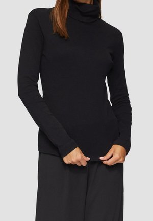 Kobieta w dopasowanym czarnym swetrze z długim rękawem i golfem, trzymająca dolny brzeg swetra obiema rękami na jednolitym jasnym tle.