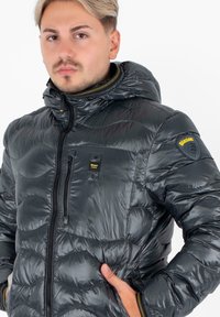 Giubbotto puffer nero con cappuccio, caratterizzato da una texture lucida, chiusura con zip e un logo giallo sul maniche. Design trapuntato per isolamento.