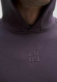 Lilla hættetrøje med blød tekstur, der har en kængurulomme og broderet "LU"-logo i matchende farve foran.