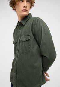 Grünes Cordhemd mit durchgehender Knopfleiste, zwei Brusttaschen und strukturiertem Stoff. Klassischer Kragen mit sichtbaren Steppnähten.