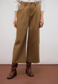Bruine wijde broek heeft twee voorsprongen aan de voorkant, knopen als sluiting en zijzakken. Gepaard met bruine enkellaarsjes voor een samenhangende look.