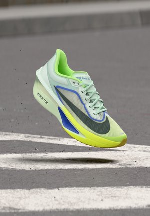 Pantof de alergare Nike Zoom Fly verde deschis și galben, în aer deasupra asfaltului cu marcaje rutiere albe.