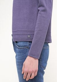Pull violet texturé avec un ourlet côtel é et un petit accent métallique. Porté avec un jean bleu, montrant une coupe ajustée et des manches longues.