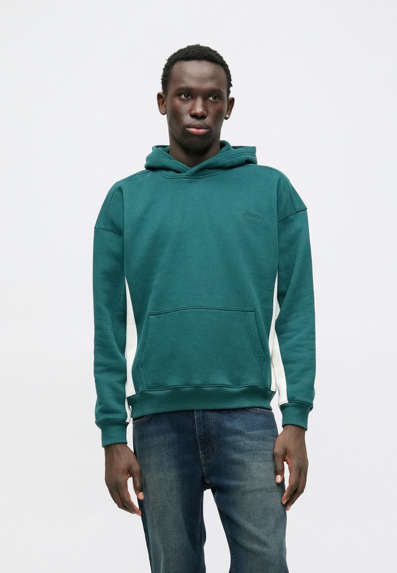 Zielona bluza z kapturem, z przednią kieszenią, białymi akcentami po bokach oraz sznurkiem do ściągania, zestawiona z niebieskimi dżinsami. Casualowy, miękki materiał.