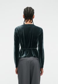 Tummansininen samettinen peplum-toppi pitkillä hihoilla, pyöreällä pääntiellä ja sidottavalla vyötäröyksityiskohdalla; pehmeä rakenne ja taitteellinen takasuunnittelu.