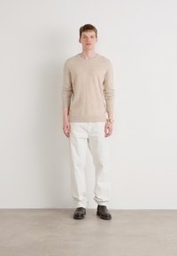 Pull beige à manches longues en matière douce, associé à un pantalon blanc. Le mannequin porte des chaussures noires et une montre en or. Tenue simple et décontractée.