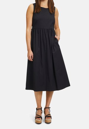 Robe noire sans manches avec un col rond, une taille froncée et des poches latérales. Personne portant des sandales à plateforme noires avec des talons en liège.