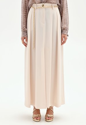 Femme portant un pantalon beige taille haute à jambes larges avec une ceinture chaîne dorée, assorti à un chemisier à manches longues imprimé léopard et des sandales dorées.