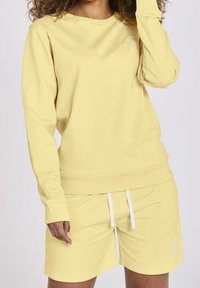 Felpa gialla con maniche lunghe e collo rotondo, abbinata a pantaloni corti coordinati. Entrambi sono realizzati in un tessuto morbido con un design minimalista.