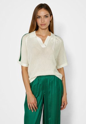 Woman with straight brown hair wears a white mesh short-sleeve polo shirt and green satin wide-leg pants, standing against a white background.