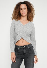 Szary dzianinowy sweter crop z prążkowanym splotem, dekoltem w kształcie litery V i kopertowym krojem, z długimi rękawami i teksturowanym wykończeniem. W zestawieniu z czarnymi dżinsami.