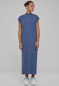 Rochie lungă, albastră, fără mâneci, dintr-un material moale, cu gât înalt și fante laterale. Asortată cu adidași albi pentru un contrast și un aspect casual.