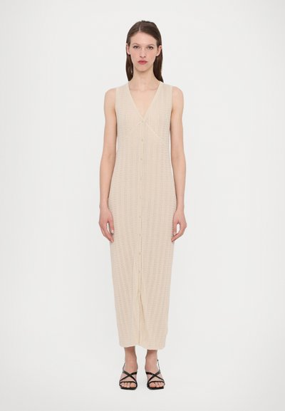 Robe midi en mailles, sans manches, décolletée en V, beige avec un devant boutonné et une texture côtelée subtile. Associée à des sandales noires à lanières.