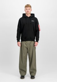 Hoodie preta com bolso frontal, capuz com cordão e um patch do logo; combinada com calças largas verdes olivas e sapatos pretos.