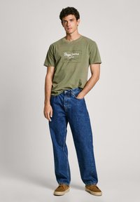 Alyvuogių žalia medvilninė marškinėliai su balta "Pepe Jeans" logotipu; derinama su laisvais mėlynais džinsais; modelis stovi vilkėdamas rudus batus.