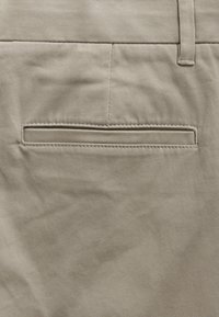 Close-up of beige fabric trousers showing a single back welt pocket and belt loop on the waistband.