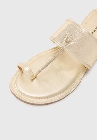Kultaiset metalliset thong-sandaalit, joissa on teksturoitu hihna, varvassuoja ja sileä pohja; ommeltu yksityiskohta reunoilla.