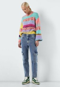 Suéter de punto multicolor con mangas anchas, con franjas horizontales en amarillo, rosa, menta y púrpura. Combinado con jeans cargo azules y zapatillas verdes.