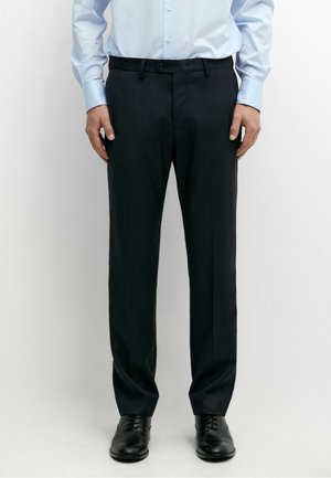Homme portant un pantalon noir formel, une chemise bleu clair et des chaussures habillées noires, debout devant un fond uni.