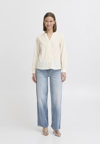 Camicia button-up beige chiaro con righe verticali, maniche lunghe e scollatura a V, abbinata a jeans a gamba larga blu chiaro e sandali argentati.
