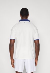 Biela polokošeľa s námorníckym golierom a manžetami. Má rebrovanú textúru a malú modrú logo na chrbte. Krátke rukávy, voľný strih.