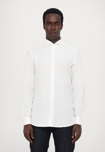 Chemise blanche à manches longues avec un col mandarin, fabriquée en tissu léger. Elle possède un devant boutonné et un ourlet légèrement arrondi. Portée avec un jean foncé.
