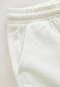 Bílé bavlněné tepláky s elastickým páskem a bočními kapsami, s jemnou texturou a viditelnými švy.